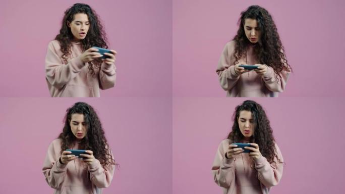 情绪化的年轻女子在粉红色背景上玩视频游戏触摸智能手机屏幕