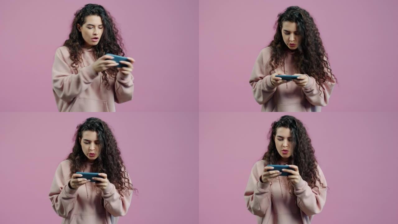 情绪化的年轻女子在粉红色背景上玩视频游戏触摸智能手机屏幕