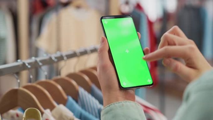 服装店: 女性使用色度键绿屏显示的智能手机。背景中带有时尚品牌商品的衣架，用于零售。特写移动设备的镜