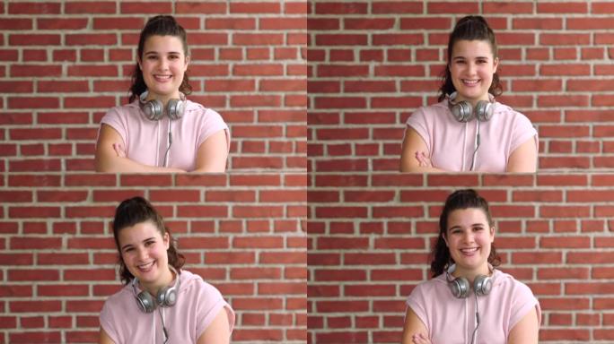 一个自信的女学生的肖像，双臂交叉站在学校的砖墙外。面对一个笑着的年轻女孩，带着耳机在户外放松，态度开
