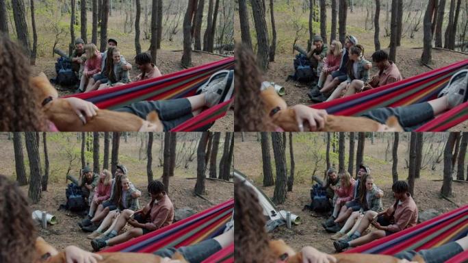 年轻女子与可爱的狗在吊床上放松，看着朋友在森林里弹吉他和唱歌