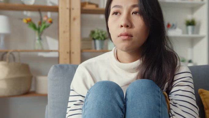 体贴的亚洲女士坐在客厅沙发上拥抱膝盖，看着外面，感到孤独，悲伤沮丧的少年独自呆在家里。