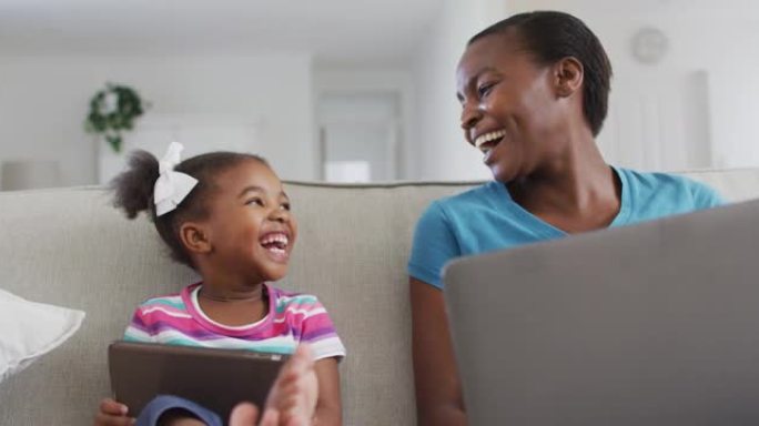 快乐的非洲裔美国母亲和女儿坐在沙发上使用数字平板电脑，笔记本电脑和笑声