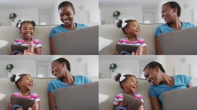 快乐的非洲裔美国母亲和女儿坐在沙发上使用数字平板电脑，笔记本电脑和笑声