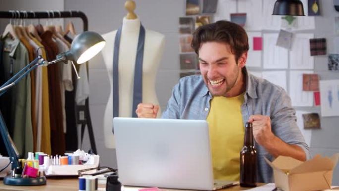 男性时装设计师带着一瓶啤酒在笔记本电脑上观看体育比赛并在工作室庆祝