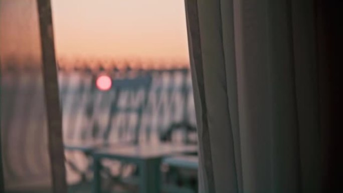 日落在海上，从酒店房间可以看到阳台和透明窗帘，椅子和桌子站在阳台上