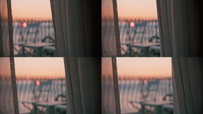 日落在海上，从酒店房间可以看到阳台和透明窗帘，椅子和桌子站在阳台上