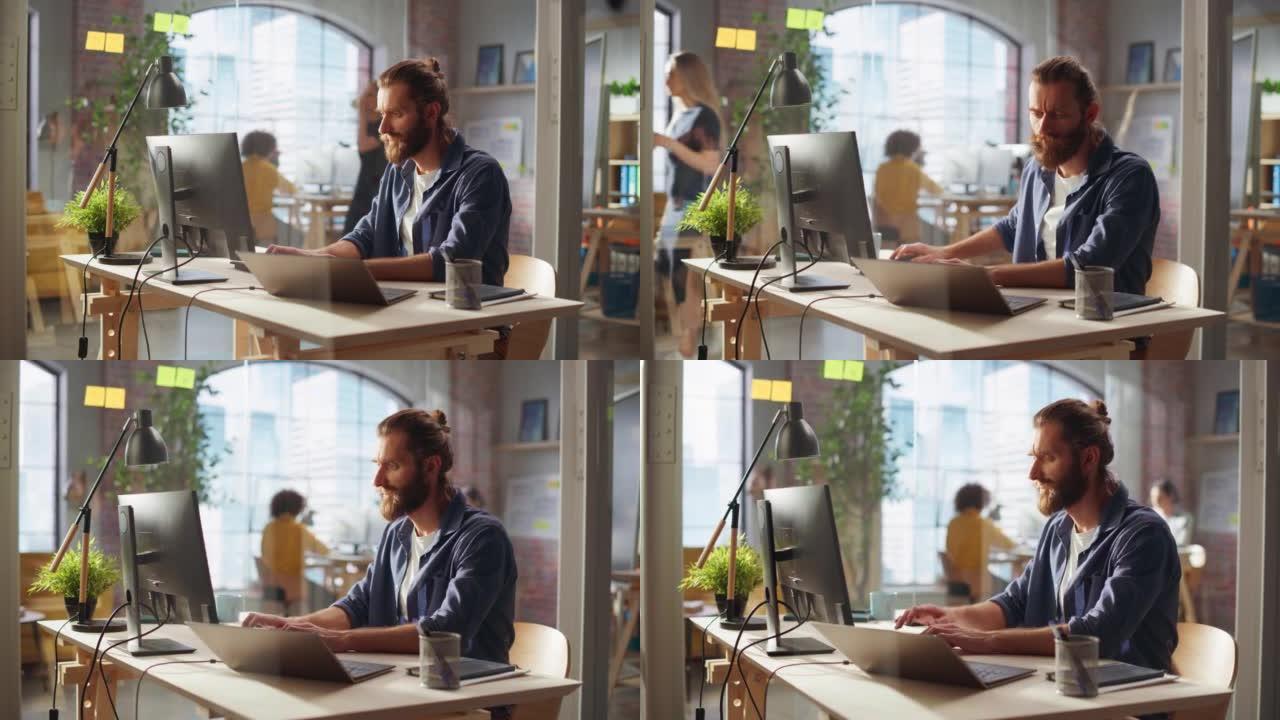 时尚的长发胡须专家坐在创意机构的办公桌前。年轻的时尚男人在连接到第二个显示器的笔记本电脑上工作。后台