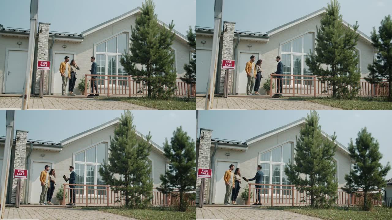 快乐的夫妇站在户外美丽的郊区房子附近与房地产经纪人交谈