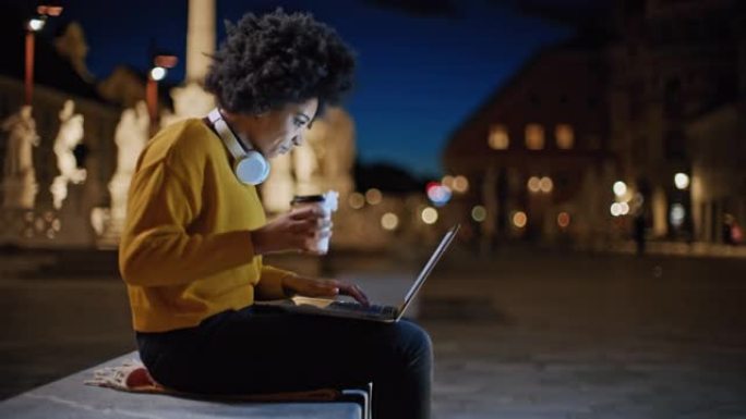 DS年轻的黑人妇女晚上在城市使用笔记本电脑时喝咖啡