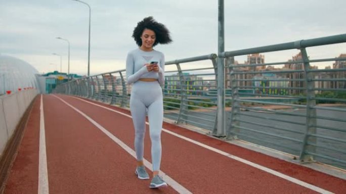 一位年轻漂亮的女跑步者穿着白色运动服，黑色卷发，正在电话里聊天，走在桥上