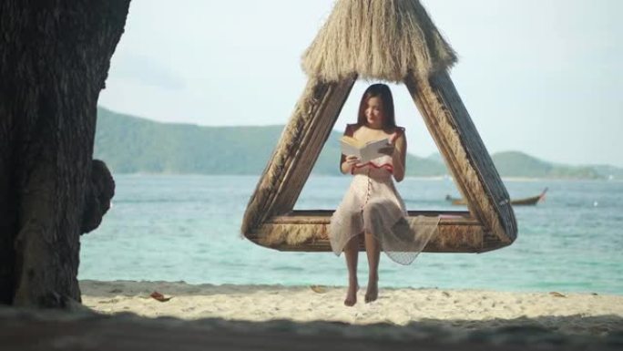 27岁的亚洲年轻女性的假期，坐在泰国普吉岛度假胜地的吊床上看书。她在海滩上享受阳光明媚的一天。泰国普