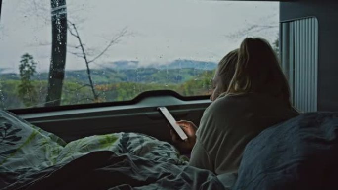 金发碧眼的女人躺在货车内的床垫上，毯子上，在下雨天用智能手机发短信