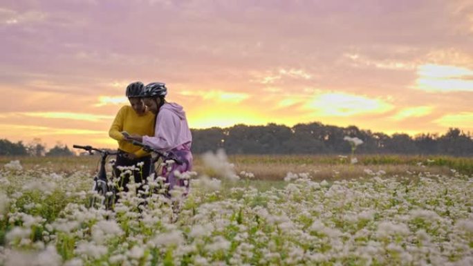 手持慢动作的两名成年女性看着智能手机，在充满花朵的草地上骑行，背景为日落