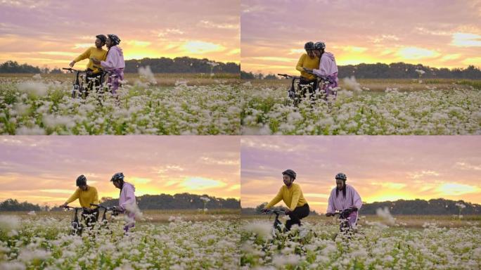 手持慢动作的两名成年女性看着智能手机，在充满花朵的草地上骑行，背景为日落