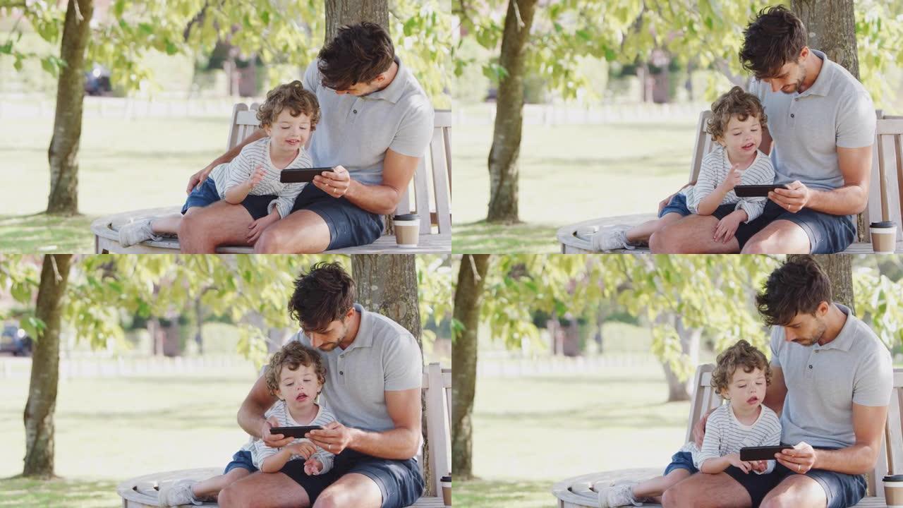 父亲坐在树下的公园长椅上，儿子一起看手机