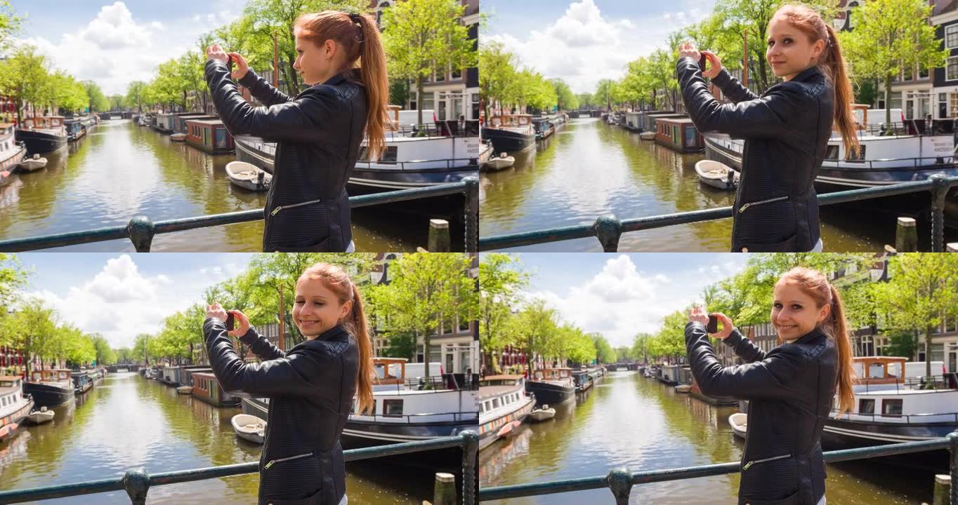 女人在阿姆斯特丹旅行，站在满是船屋的运河上的桥上，用智能手机拍照