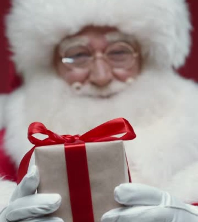 一个高兴的圣诞老人的特写镜头，微笑着向相机展示一个可爱的小礼品盒，站在红色背景上