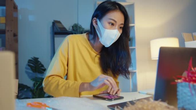 年轻的亚洲女商人戴着口罩检查智能手机上的采购订单，晚上在家庭办公室的电脑中库存。