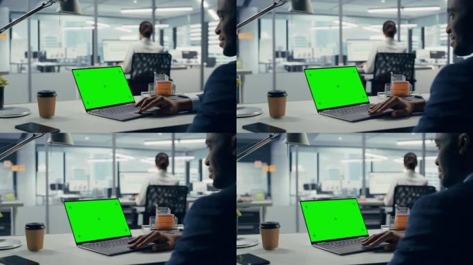 成功的黑人商人坐在办公桌前，在大城市办公室工作绿屏色度钥匙笔记本电脑。努力工作的高级经理为电子商务项