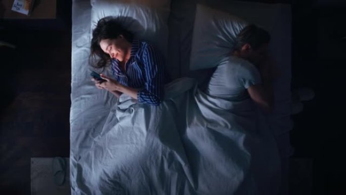 顶视图: 年轻女子晚上在床上使用智能手机，当时她的女性伴侣试图在旁边入睡。夫妻吵架，争论。社交媒体上