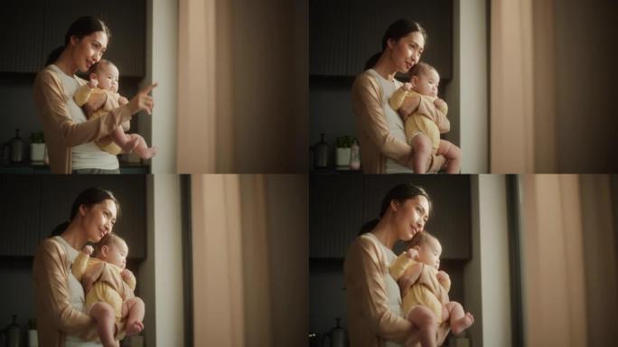 美丽的年轻亚洲母亲抱着婴儿，站在家里的窗户旁边。快乐的女人，对母亲的新手，拥抱她的孩子，一起享受亲密