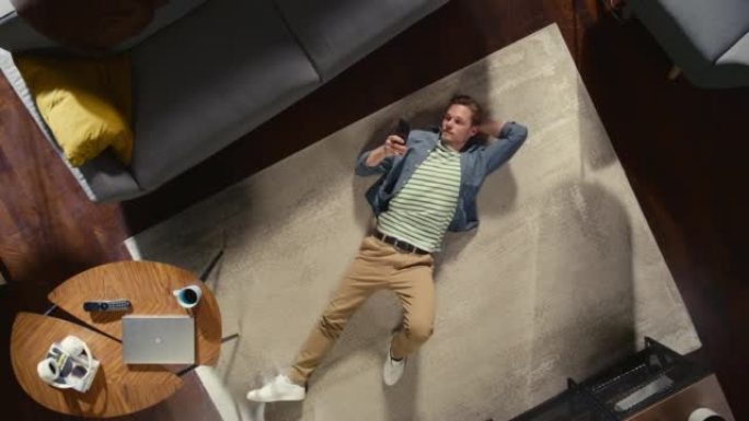 公寓:年轻人躺在客厅的地板上，用智能手机。盖伊在Сarpet上放松。自由职业者远程工作，电子商务，网