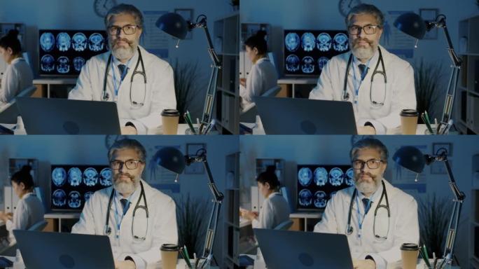 经验丰富的男医生的肖像坐在桌子旁，晚上在医院用电脑看着相机