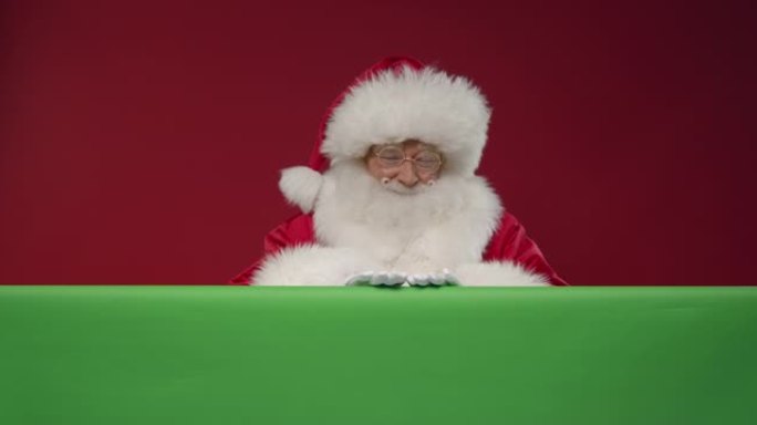 圣诞老人从他面前的绿色屏幕后面出现在红色背景的框架中，用手仔细触摸屏幕，竖起大拇指并走开