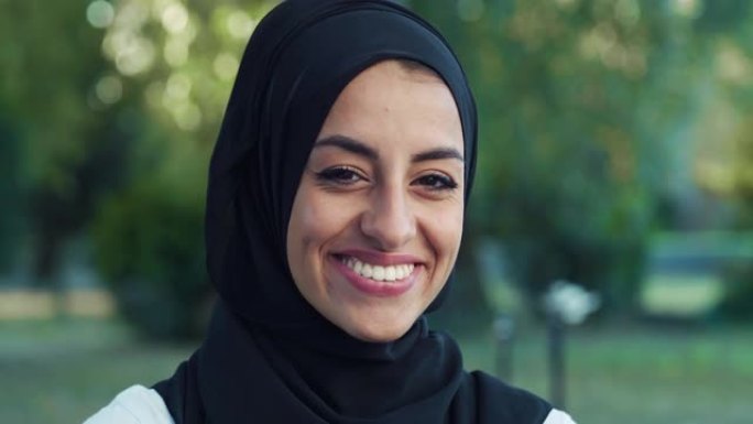 特写美丽的年轻阿拉伯女人看着相机，带着绿色背景微笑的肖像。戴着头巾的穆斯林女少年在公园里享受新鲜空气
