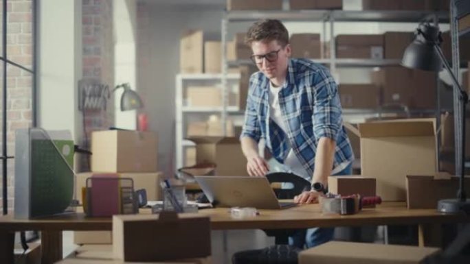 小企业主站在仓库的办公桌前，在笔记本电脑上工作。员工在房间里用装满纸板箱包裹的架子包装一个时尚的复古