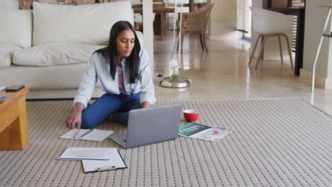混合种族妇女使用笔记本电脑通过文书工作在家喝咖啡