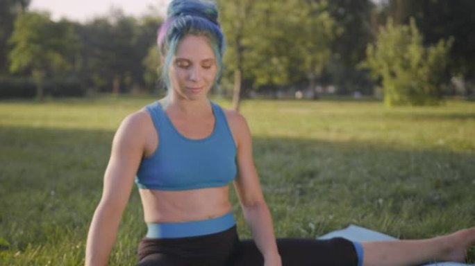 迷人的年轻女子，蓝色的头发，穿着蓝色的瑜伽服，在公园的草地上做瑜伽，在日出或日落时，通过左右转动来伸