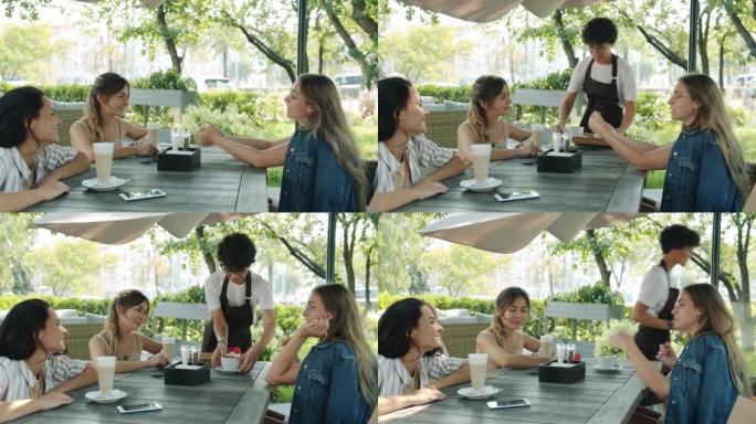 快乐的女性朋友在露天咖啡馆聊天和大笑，而围裙的女服务员在夏日带食物和饮料
