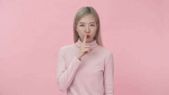 工作室的肖像年轻美丽的亚洲妇女在粉红色的长袖t恤举起手指到嘴唇显示沉默或安静的标志在粉红色的背景相机