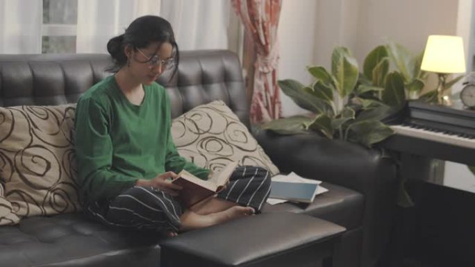手持镜头: 亚洲少女呆在家里读一本模拟书。她翻开学习书的一页。亚洲学生在她家的学校学习历史课作业。