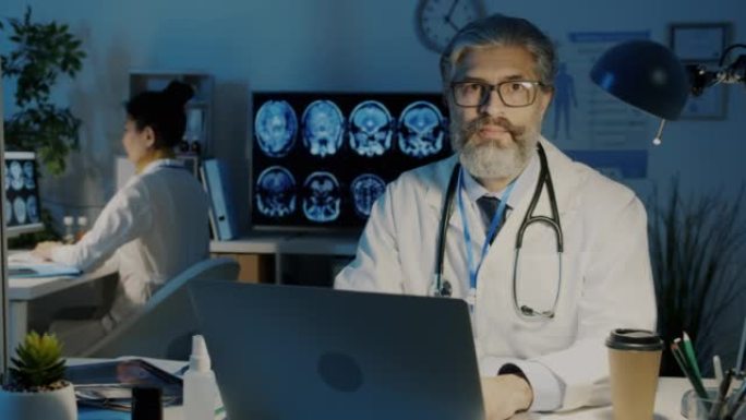 穿着白色礼服的经验丰富的医生的慢动作肖像坐在办公桌前，笔记本电脑在黑暗的办公室