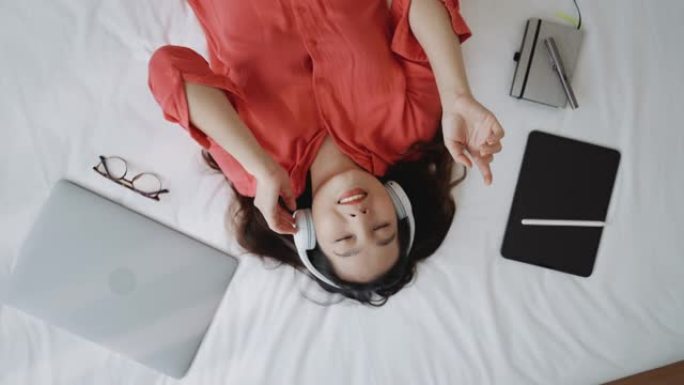 戴着耳机的亚洲妇女在家中躺在床上使用智能手机