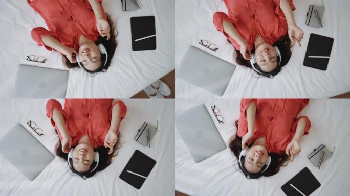 戴着耳机的亚洲妇女在家中躺在床上使用智能手机