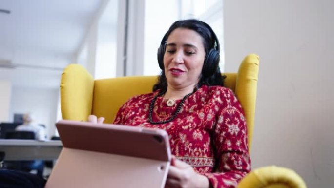 成熟的女人，耳机坐在扶手椅上，在办公室使用数字平板电脑