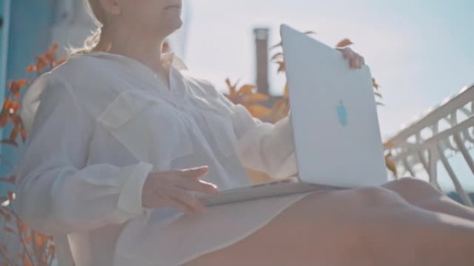 金发碧眼的女人坐在酒店房间的阳台上，天气晴朗，阳光直射，停止在笔记本电脑上工作