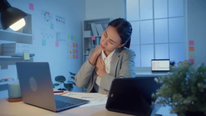 年轻的亚洲女商人坐在桌子上，用笔记本电脑和平板电脑摩擦眼睛，晚上在办公室因劳累而感到疼痛和疲倦。