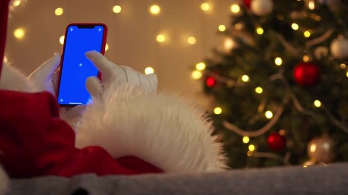 圣诞老人使用带有蓝色色度键的智能手机，我们从后面看到他坐在圣诞树前