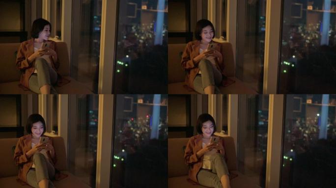 亚洲中国美女晚上在沙发上的客厅用手机靠近城市的窗户