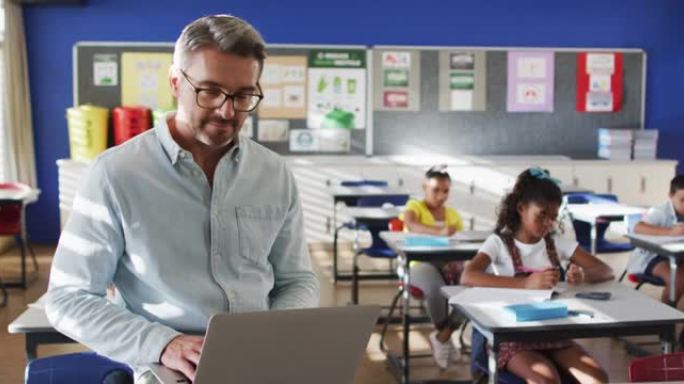 快乐的高加索男老师在教室里的肖像，孩子们用笔记本电脑看着相机