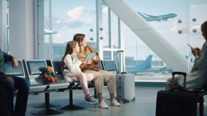 繁忙的机场航站楼: 可爱的母亲和小女儿等待他们的假期航班，在数字平板电脑上玩教育游戏。坐在飞机飞行的