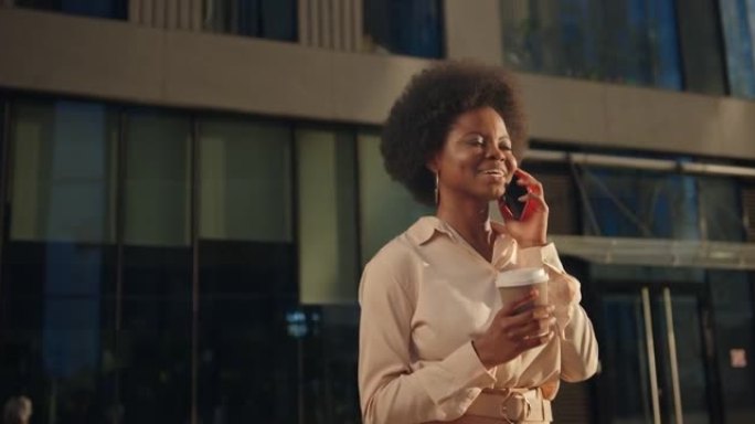 一位穿着米色服装的年轻黑皮肤妇女在现代立面的街道上行走，用手机聊天，喝咖啡