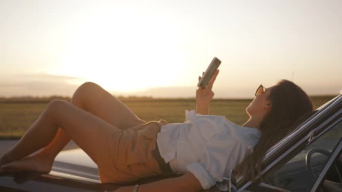 SLO MO年轻女子躺在汽车引擎盖上时使用智能手机