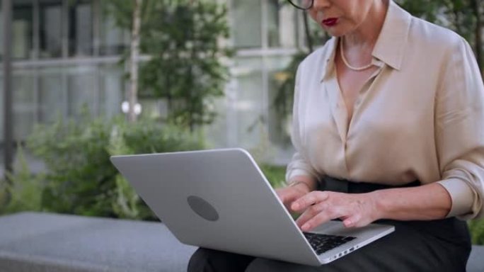 一位成功的白发女作家正坐在现代商业中心前，用笔记本电脑写一本新书，