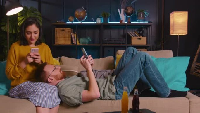 拖延概念。年轻幸福的夫妇坐着，躺在家里的沙发上放松，用手机玩手机游戏。